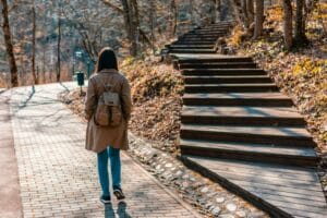 A woman in a brown coat walking through a leaf-covered park in fall takes a flat, paved path that branches from a path of stairs leading up a hill